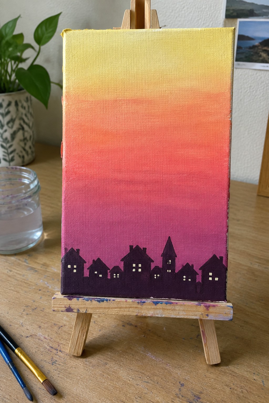 Canvas on wooden easel with yellow-to-purple sunset gradient and purple house silhouettes.