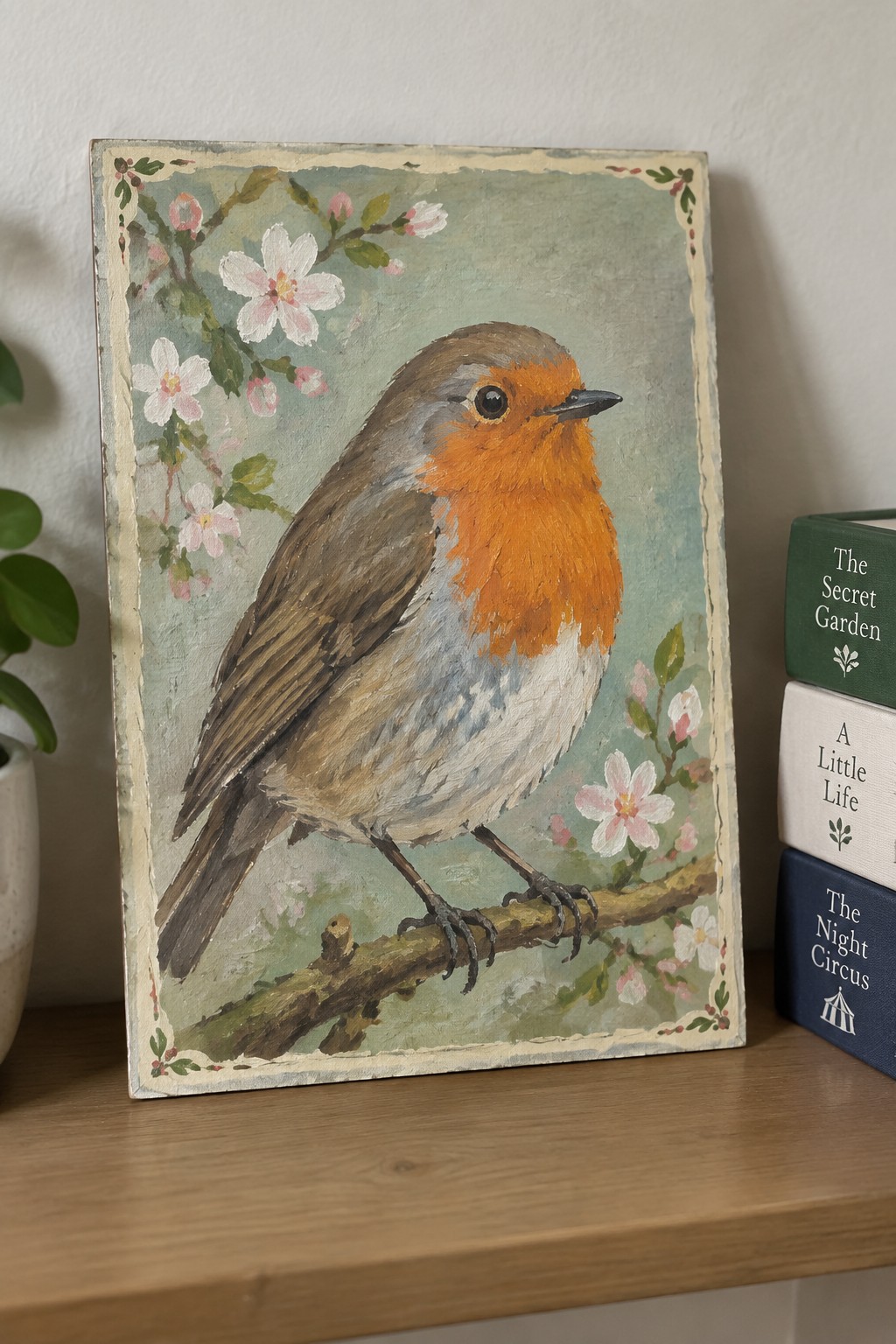 Painting of robin with orange chest on branch amid pink flowers next to books