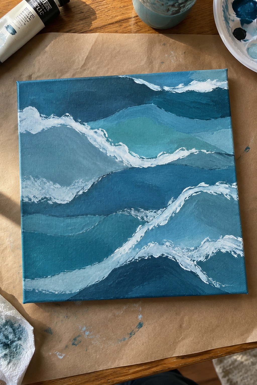 Acrylic painting on canvas showing overlapping ocean wave shapes in blue and teal with white highlights, placed on brown paper with paint supplies nearby.