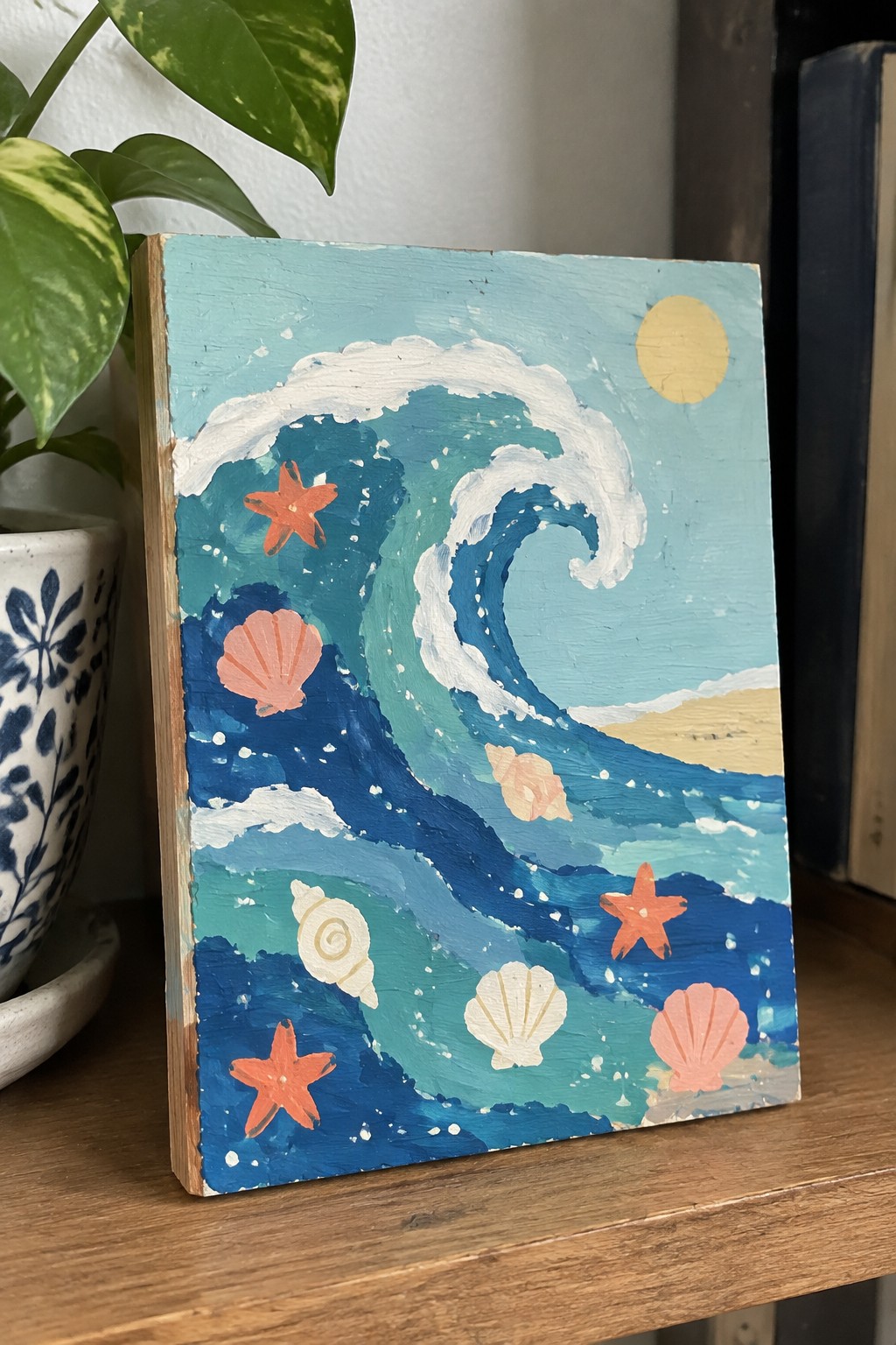 Acrylic painting of a large curling blue wave with white foam, orange starfish, and pink and white seashells on a canvas with a yellow sun and sand in the background.