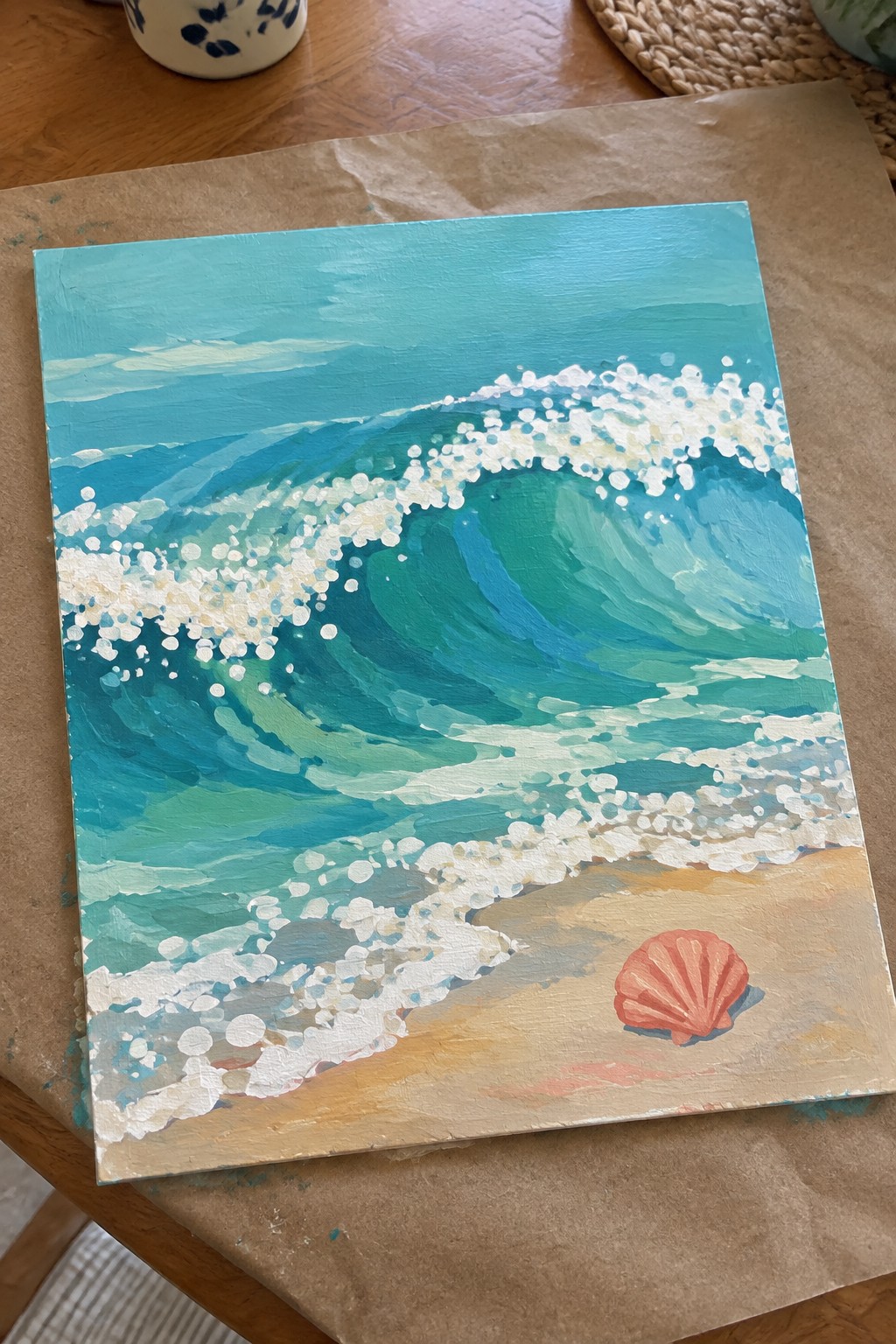 Seascape painting of curling teal wave with white foam on sandy beach beside orange shell