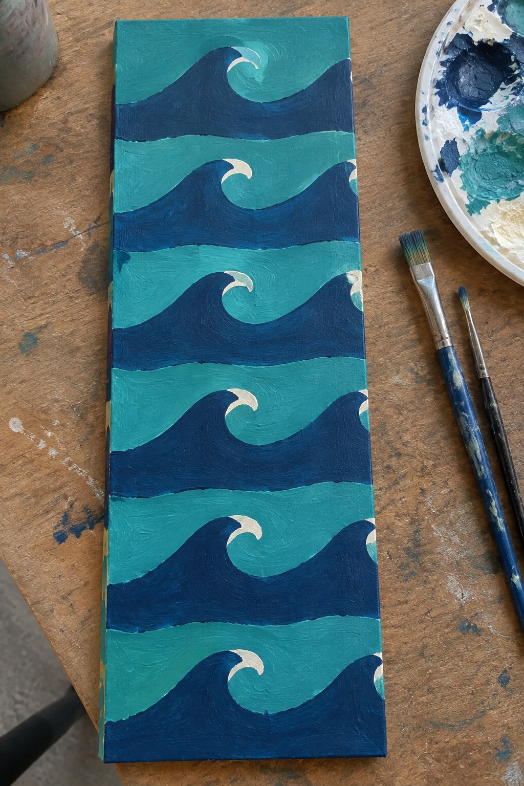 Narrow vertical canvas with blue teal wave painting beside brushes and palette on wooden table.
