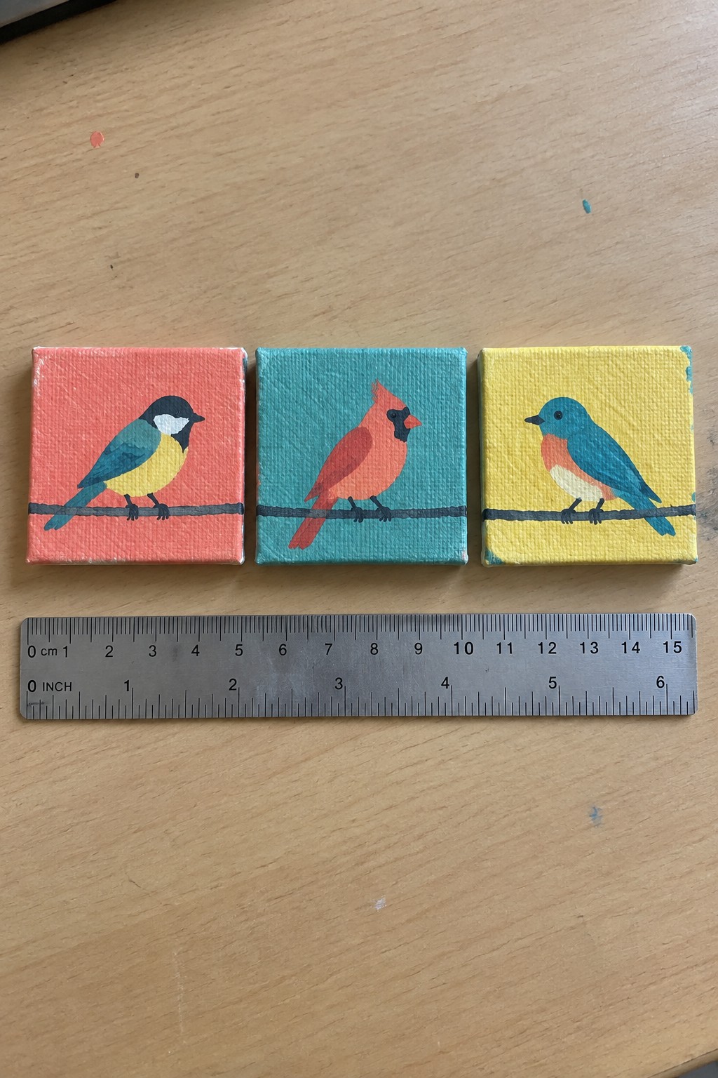Three mini canvases with colorful birds above a ruler on wood.