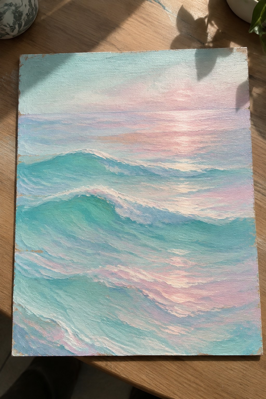 Painting of turquoise waves under soft pink and blue sky on wooden table