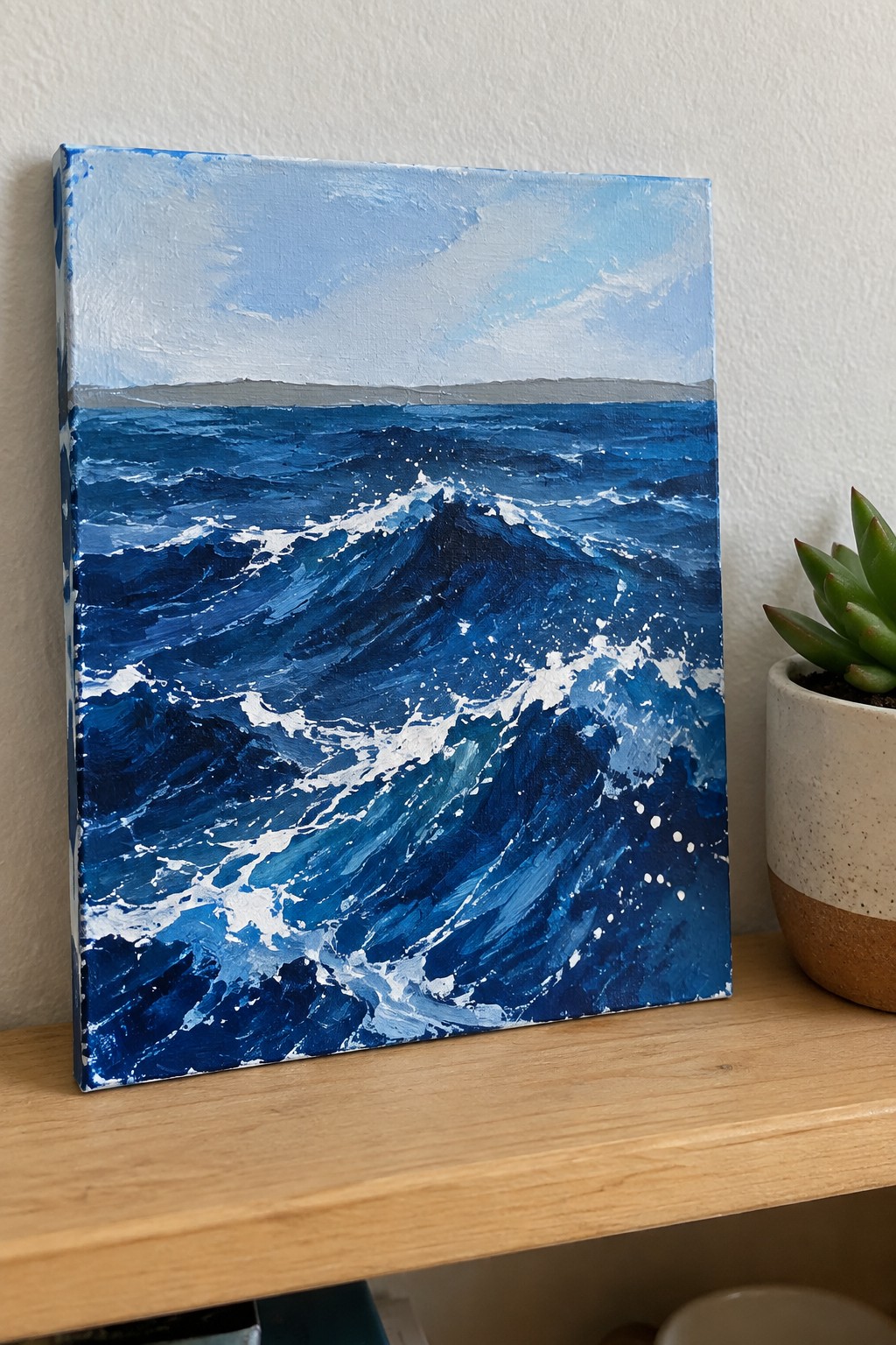 Seascape painting of crashing blue waves with white foam beside potted succulent on shelf