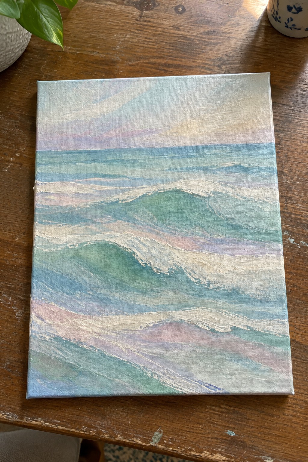 Pastel seascape painting of rolling turquoise waves on wooden table.