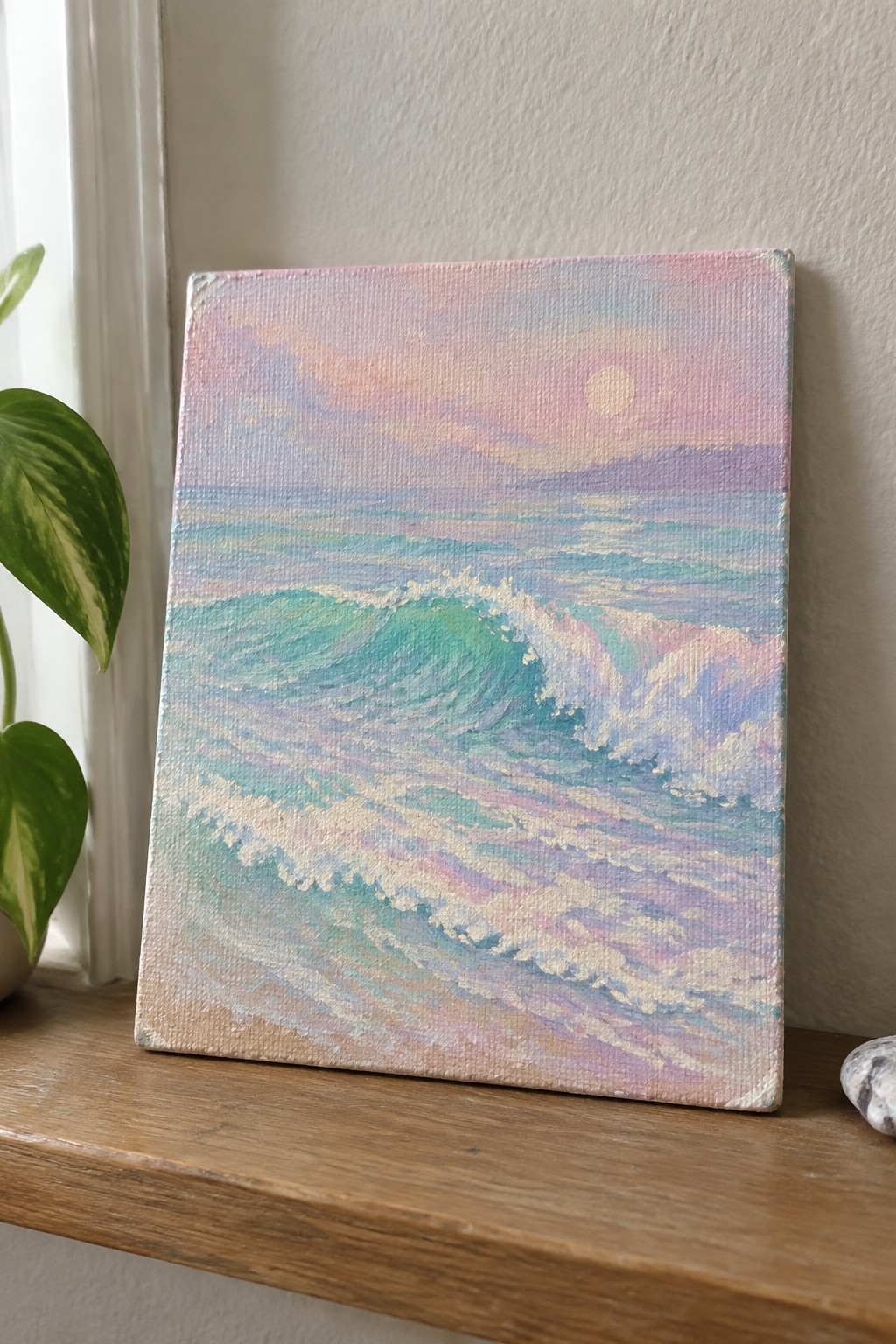 Pastel seascape painting of turquoise waves under pink sky on wooden shelf beside plant.
