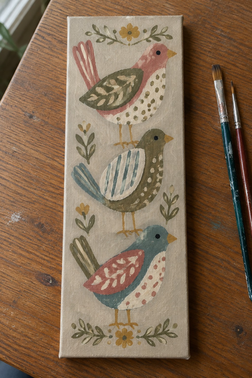 Three folk-art birds on canvas with paintbrushes on wooden table